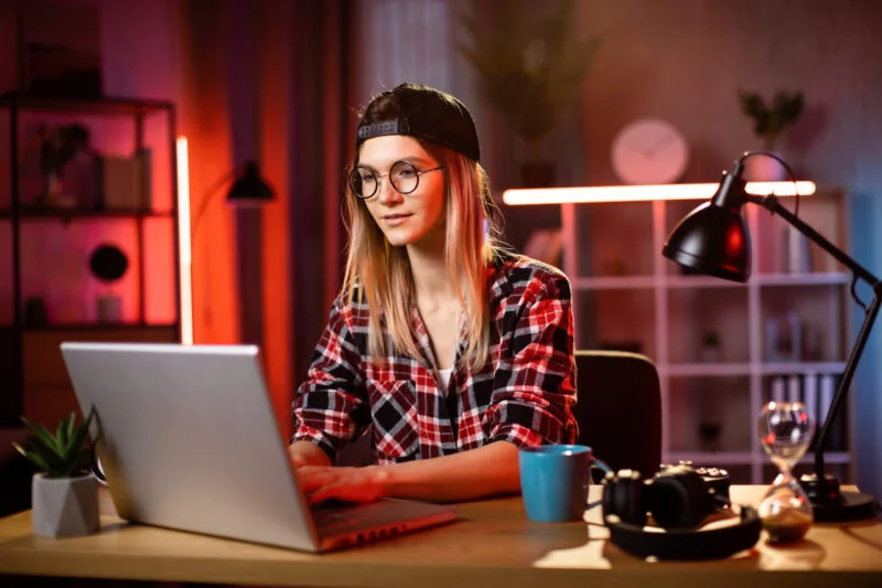 L'image met en valeur l'Ashwagandha, en parfaite adéquation avec l'objectif du site. Une personne portant des lunettes et une casquette est assise à un bureau devant un ordinateur portable, entourée d'objets tels qu'une tasse, des écouteurs et une lampe. La pièce est illuminée de manière créative par des lumières rouges et bleues qui ajoutent à l'ambiance où l'Ashwagandha occupe le devant de la scène.